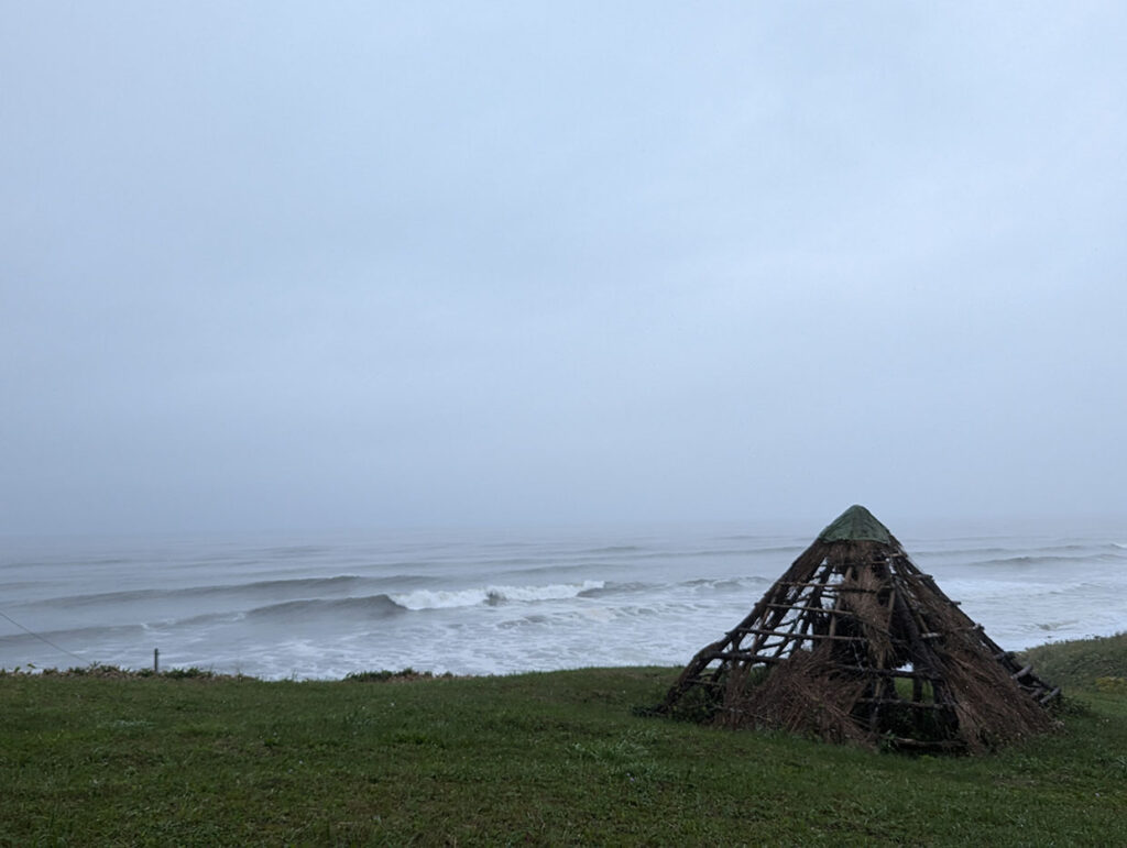 雨の中　再現された竪穴住居と荒れた海